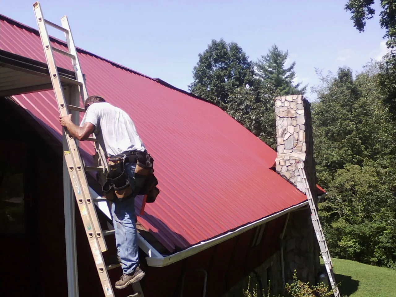 Roofer on a ladder beside a red metal roof and stone chimney — Soffit & Fascia Repair by Roofing Brothers in Rapids City